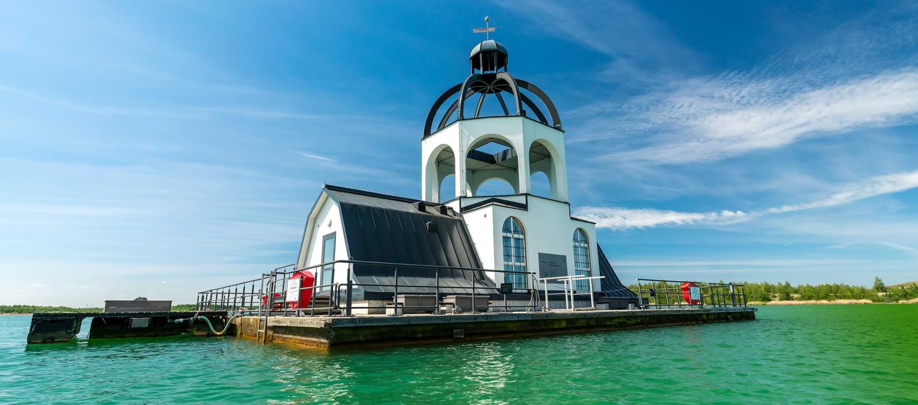 Eine kleine weiße Kapelle mit einem schwarzen Dach steht auf einem Steg inmitten von grünem Wasser. Der Himmel ist blau mit einigen Wolken. Die Architektur der Kapelle hat einen auffälligen, kuppelartigen Aufbau.