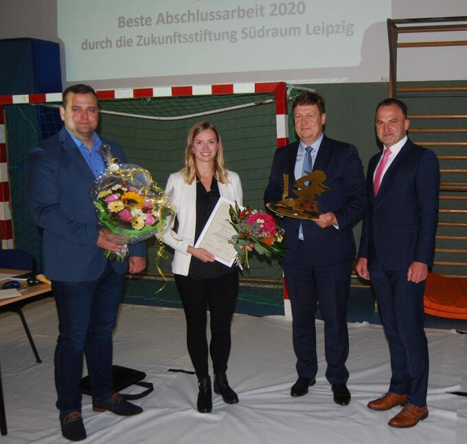 Vier Personen stehen in einem Raum. Eine Frau in der Mitte hält eine Urkunde und Blumen. Neben ihr stehen drei Männer, von denen einer einen Preis in der Hand hält. Im Hintergrund ist ein Sportfeld und ein Banner sichtbar.
