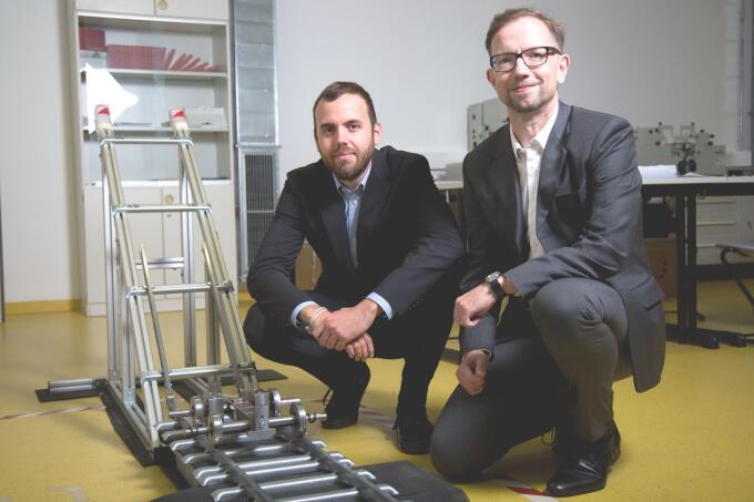 Zwei Männer in formeller Kleidung knien neben einer technischen Vorrichtung auf einem gelben Boden. Die Vorrichtung besteht aus Metall und hat Schienen und verschiedene mechanische Teile.