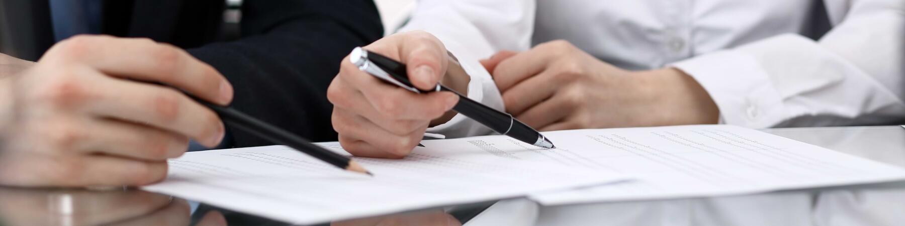 Zwei Personen sitzen an einem Tisch und prüfen Dokumente. Eine Person hält einen Stift, während die andere mit einem Bleistift auf die Papiere zeigt. Die Szene vermittelt eine geschäftliche Diskussion oder Zusammenarbeit.