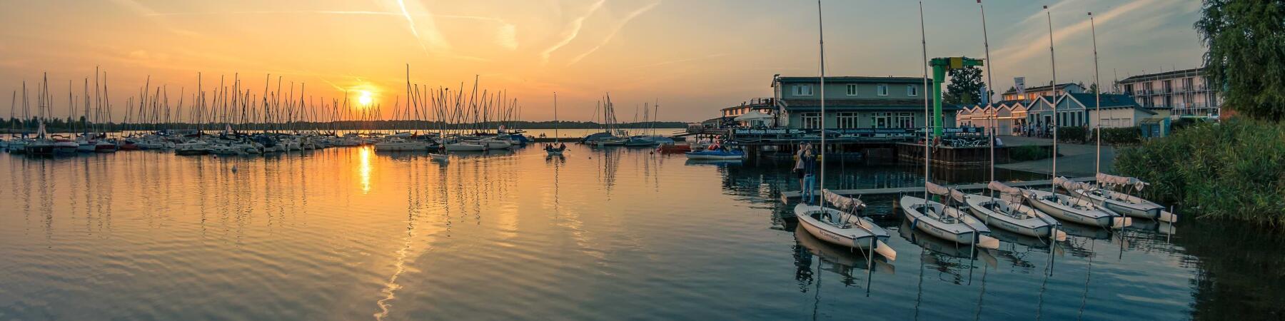 Ein Hafen am Cospudener See bei Sonnenuntergang, mit vielen Segelbooten im Wasser. Die Sonne spiegelt sich im ruhigen Wasser, während der Himmel in sanften Farben leuchtet. Am Ufer sind Gebäude und weitere Boote sichtbar, umgeben von grünen Pflanzen.