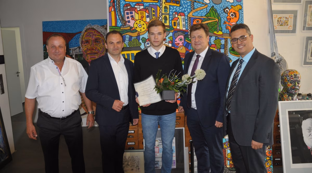 Eine Gruppe von fünf Männern steht in einem Raum mit bunten Kunstwerken im Hintergrund. Ein junger Mann in der Mitte hält ein Dokument und einen Blumenstrauß. Die anderen Männer lächeln und scheinen ihm zu gratulieren.