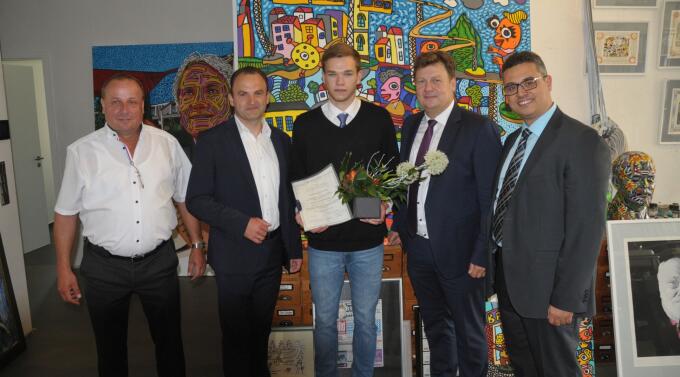 Eine Gruppe von fünf Männern steht in einem Raum mit bunten Kunstwerken im Hintergrund. Ein junger Mann in der Mitte hält ein Dokument und einen Blumenstrauß. Die anderen Männer lächeln und scheinen ihm zu gratulieren.