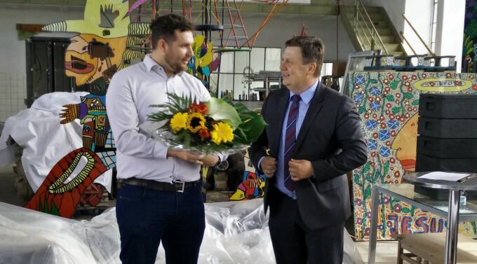 Zwei Personen stehen in einem Innenraum, einer hält einen Blumenstrauß. Sie scheinen miteinander zu sprechen. Im Hintergrund sind bunte Kunstwerke und Materialien zu sehen. Der Raum hat eine kreative, künstlerische Atmosphäre.