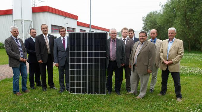 Eine Gruppe von Männern steht im Freien vor einem Solarpanel. Sie tragen formelle Kleidung und lächeln in die Kamera. Im Hintergrund sind Gebäude und Bäume zu erkennen. Das Bild vermittelt eine positive Atmosphäre im Kontext erneuerbarer Energien.