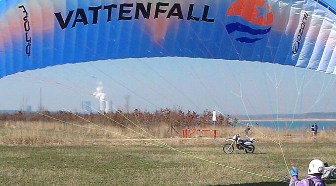 Ein Gleitschirm mit der Aufschrift "Vattenfall" schwebt über einem grünen Feld. Im Hintergrund sind das Wasser eines Sees und eine Industrieanlage sichtbar. Eine Person auf einem Motorrad ist im Vordergrund zu sehen.