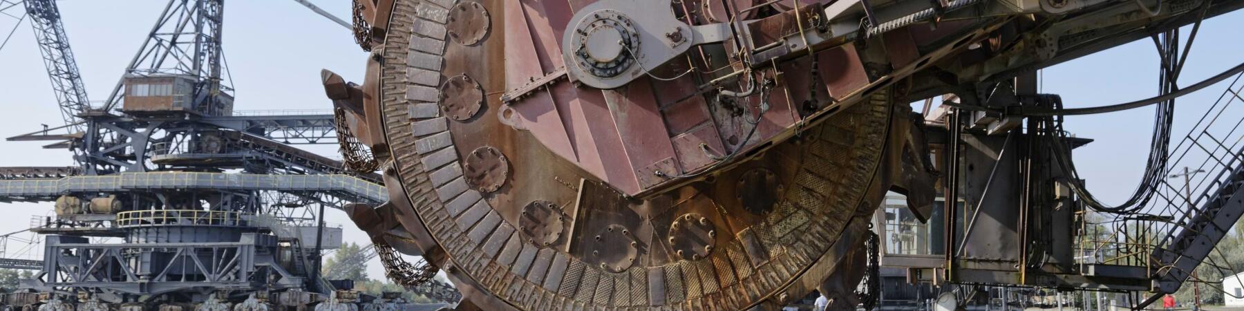 Auf dem Bild sind große, rustikale Bagger zu sehen, die in einem Freiluftbereich stehen. Die Maschinen haben massive Zahnräder und sind mit verschiedenen mechanischen Teilen ausgestattet, die für den Abbau verwendet werden. Der Himmel ist klar und blau im Hintergrund.
