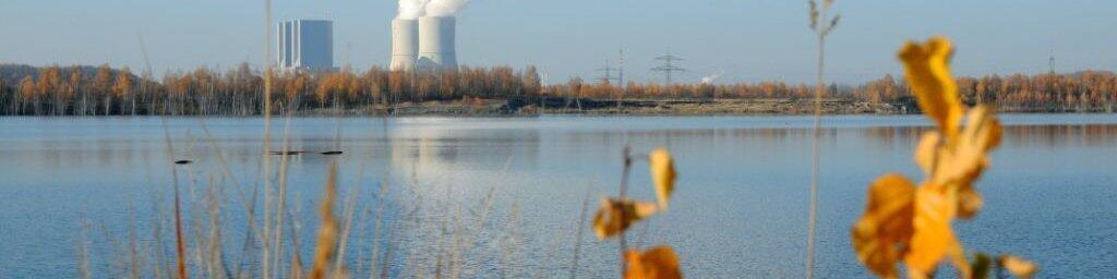 Im Vordergrund sieht man gelbe Laubblätter, während im Hintergrund ein stiller See zu sehen ist. Über dem Wasser steigen Rauchwolken aus zwei Kühltürmen eines Kraftwerks auf. Die Umgebung ist herbstlich und es gibt klare, blauen Himmel.