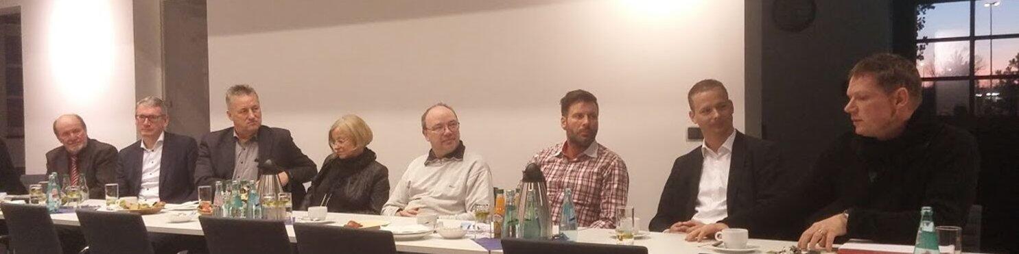 Eine Gruppe von acht Personen sitzt an einem langen Tisch in einem modernen Raum. Die Anwesenden sind in formeller Kleidung gekleidet und diskutieren; auf dem Tisch stehen Getränke und Snacks. Im Hintergrund ist eine Wand mit Fenstern zu sehen.