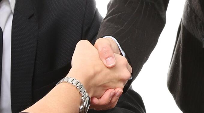 Eine Geschäftsszene mit fünf Personen, die an einem Tisch sitzen. Zwei Frauen stehen, eine reicht der anderen die Hand zur Begrüßung. Die Männer im Hintergrund beobachten das Geschehen, während auf dem Tisch ein Laptop und einige Dokumente liegen. Alle sind professionell gekleidet.